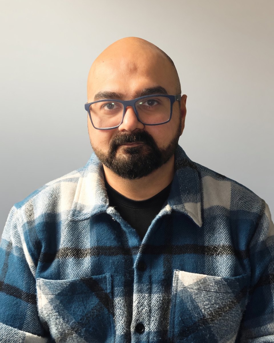 Qambar Raza, wearing blue glasses and a blue checked shirt, against a plain grey wall
